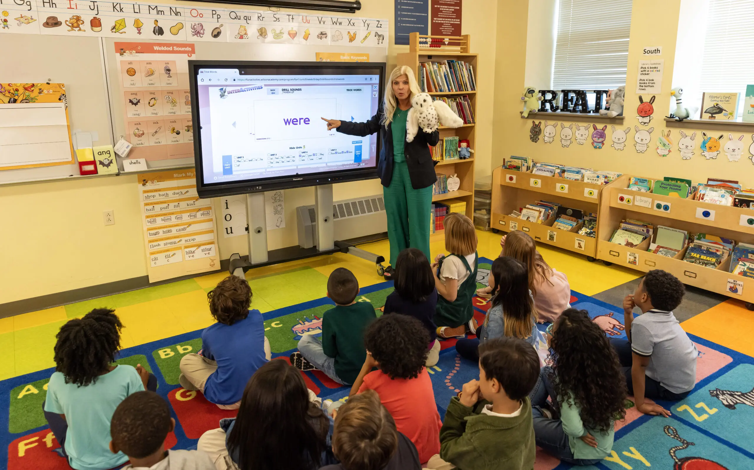 A teacher stands at the front of a classroom, pointing at a large interactive screen displaying the word were. She holds a white puppet while students sit on a colorful alphabet rug, attentively listening. Classroom bookshelves and educational posters are visible in the background.
